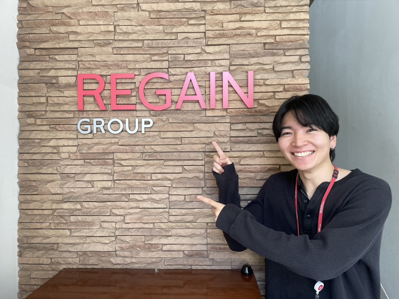 REGAIN GROUPのロゴがある壁の前でポーズをとる豊川さん