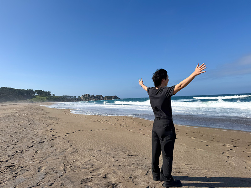 青空の下、青森の海岸で両手を広げる豊川さん
